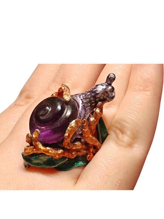 Close-up of the Amethyst Wanderer ring worn on a hand from the Stone Garden Collection by Konforti Jewelry, handcrafted in 960 silver and set with a 29 ct lab-grown amethyst carved into a spiral snail shell, detailed with hand-painted Limoges-style enamel in vivid layered colors.