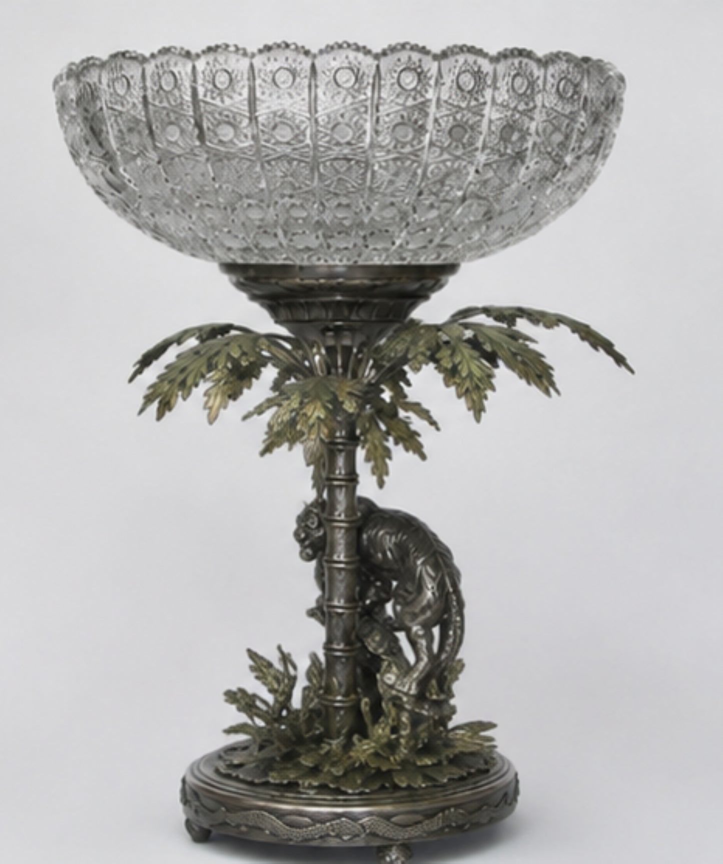 An ornate pedestal bowl featuring a clear cut-glass bowl mounted on a bronze and green metal base sculpted with a palm tree and a lion.