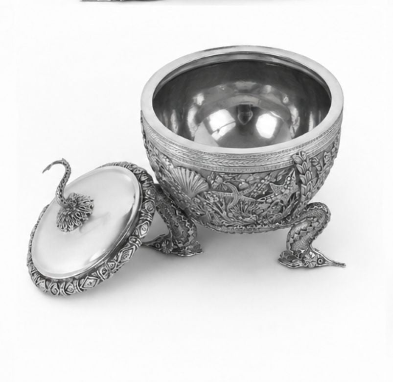 An ornate silver-toned metal bowl with a matching lid, featuring detailed floral and scrollwork engravings and three sculpted legs, photographed on a plain white background.