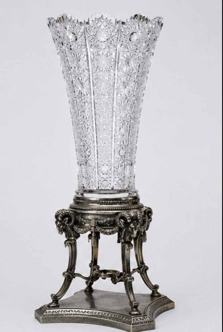 A clear cut-glass or crystal vase with a flared, ornate rim, displayed on an ornate silver pedestal stand against a light gray studio background.