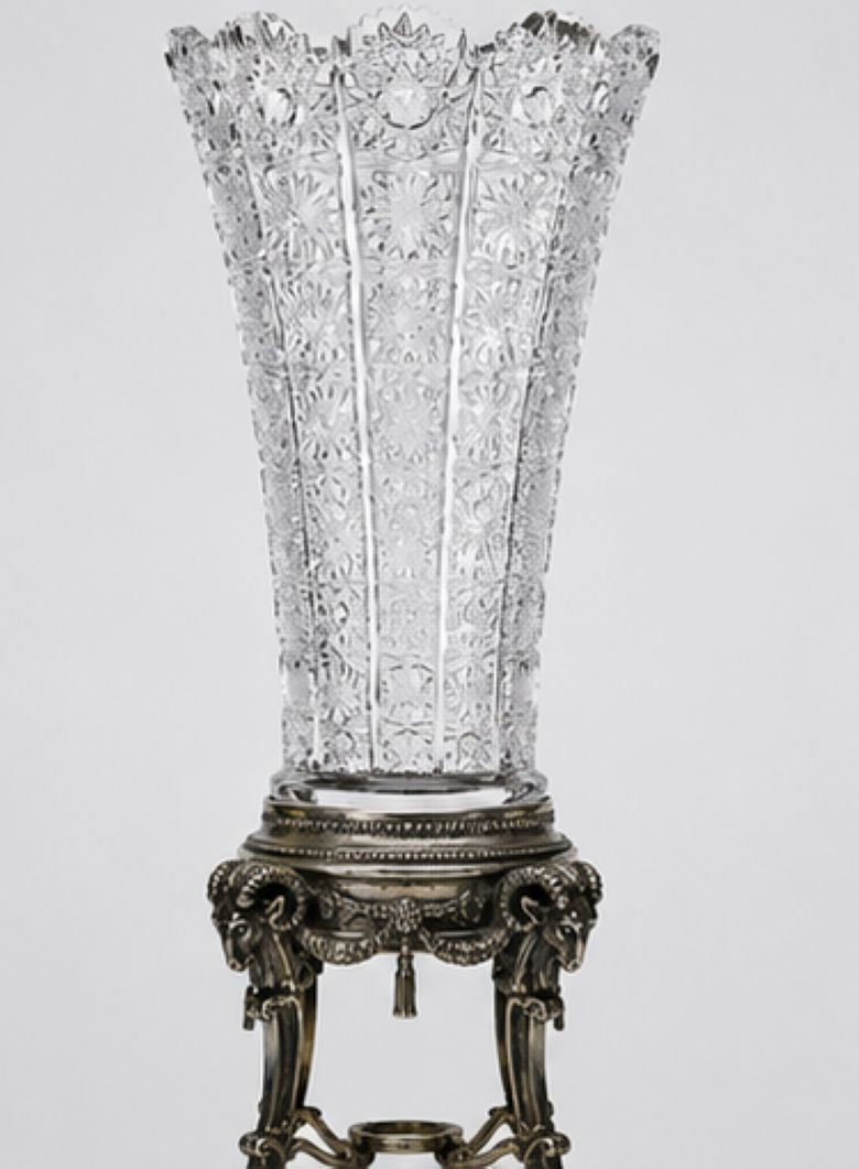 A clear cut-glass vase with a wide, flared rim and ornate vertical facets, displayed on an antique silver-toned pedestal stand against a light gray background.
