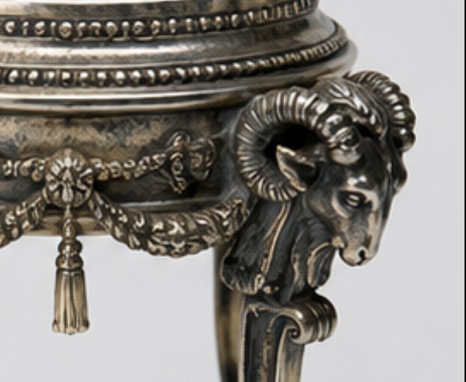 Close-up of an ornate antique-style metal stand or pedestal featuring a ram head leg and decorative beaded trim.