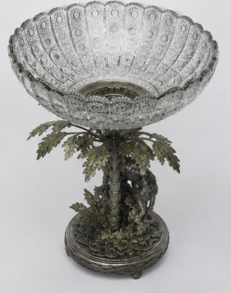 A decorative pedestal bowl with a clear cut-glass or crystal upper bowl mounted on an ornate silver-toned metal base shaped like a palm tree with a horse and foliage, photographed against a plain light background.