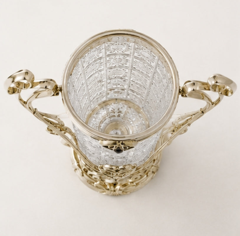 An ornate silver-toned metal cup with a clear cut-glass body, featuring decorative handles and a patterned interior, photographed on a plain light background.