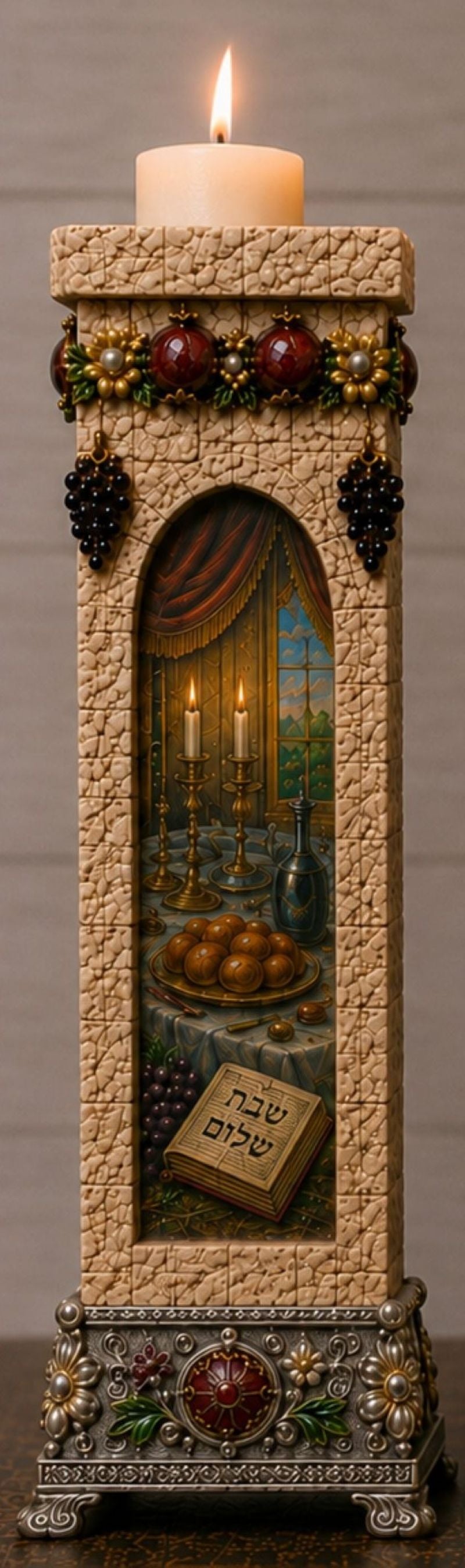 A tall decorative candle holder with a lit white candle on top, featuring a textured beige stone-like body, an arched inset scene with a table, wine, and a book with Hebrew text, and red and green decorative accents around the top and base.