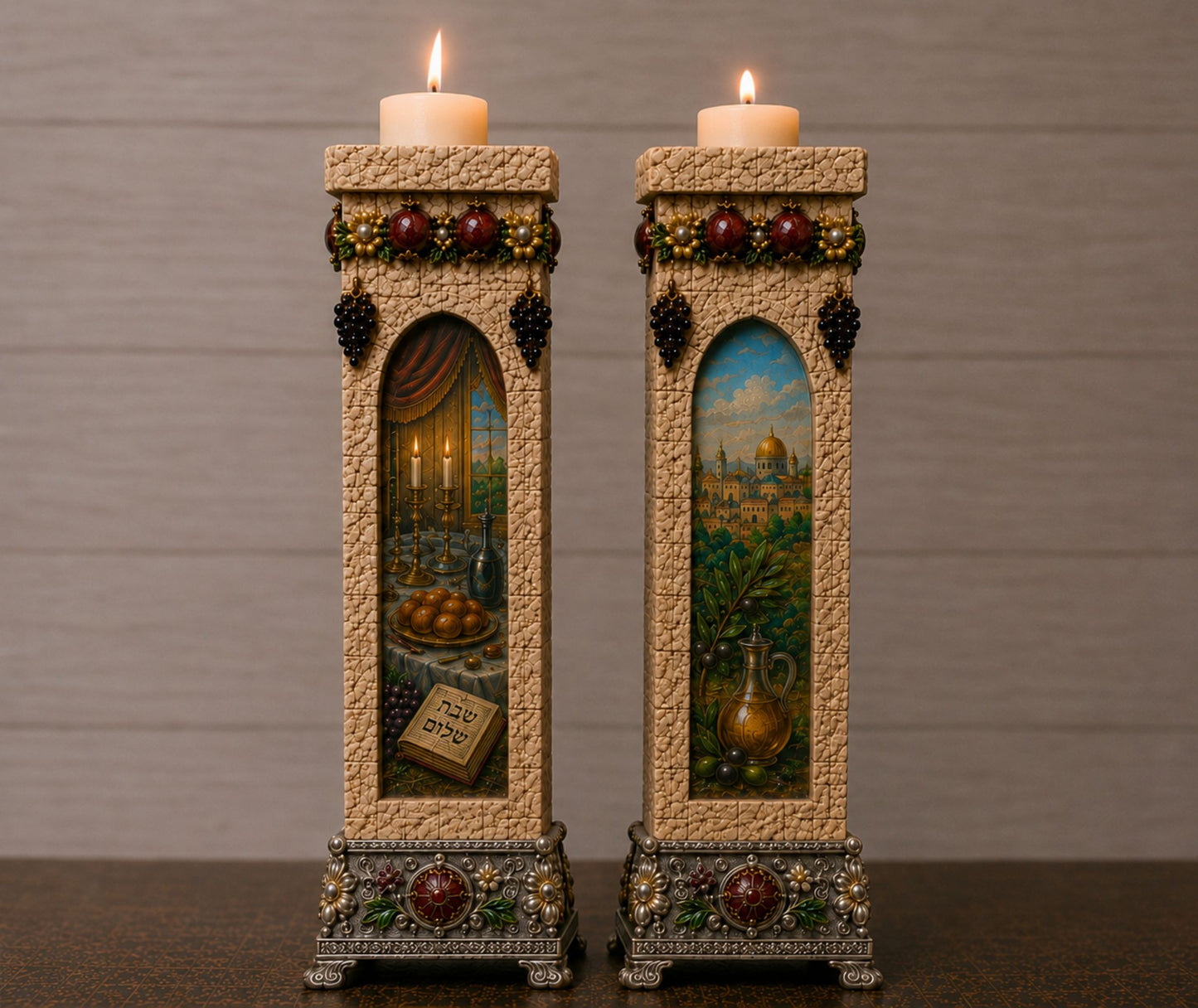 Two tall decorative candle holders with lit candles, featuring detailed painted scenes and ornate metallic bases on a dark tabletop against a gray paneled background.