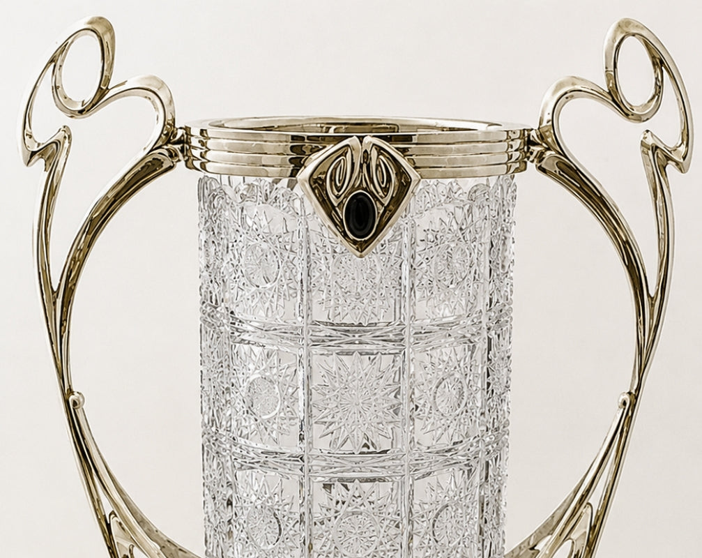 A close-up of a clear cut-glass cylinder container with ornate silver-toned metal handles and a decorative crest at the rim, shown against a plain light background.