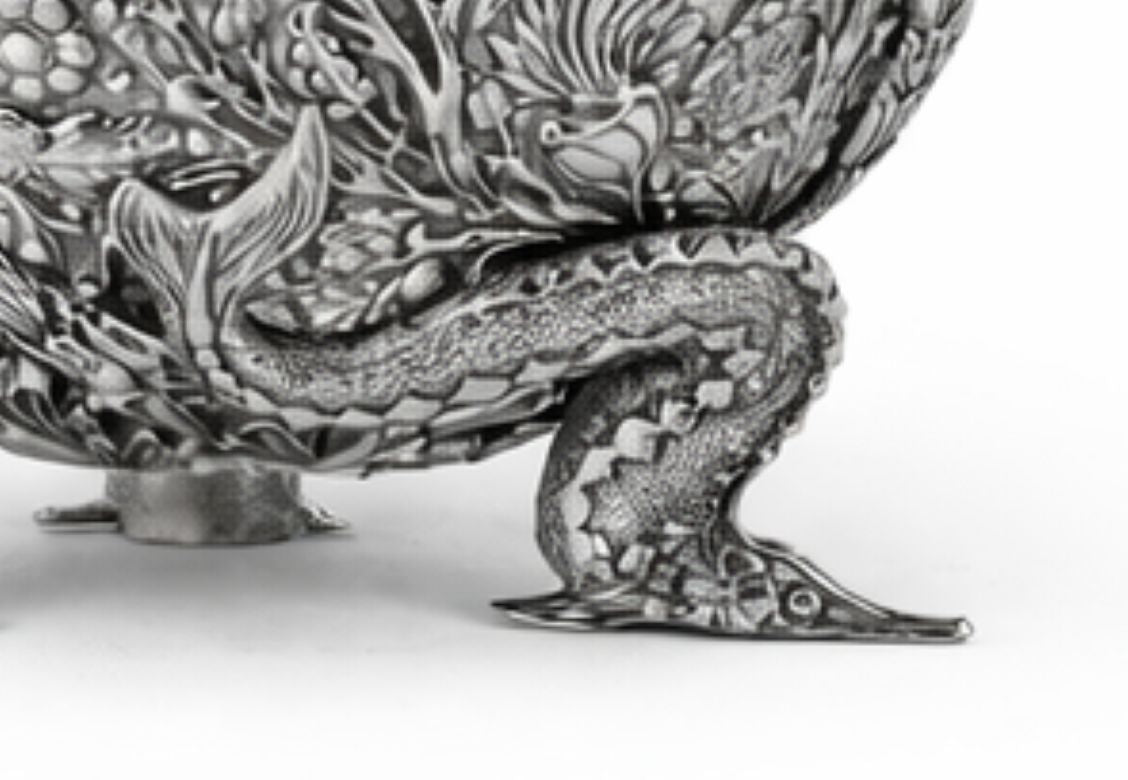 Close-up of an ornate silver-toned metal object featuring intricate dragon or serpent-like sculpted details and a curved clawed foot, photographed against a plain white background.