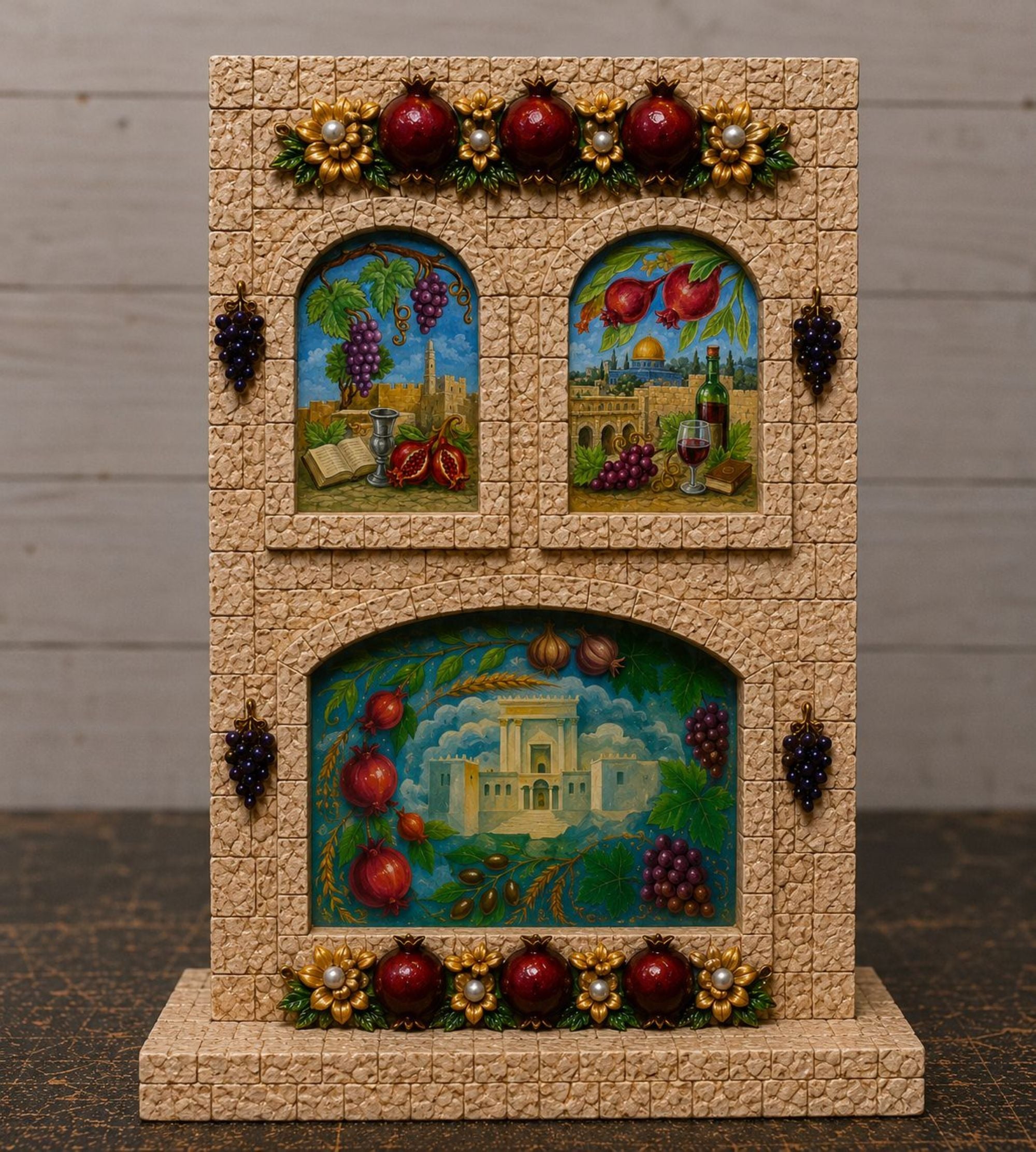 A decorative wine-themed tabletop display featuring a textured beige stone facade with two arched panels and a larger lower panel. The panels depict grapes, wine bottles, a wine glass, and a vineyard scene.
