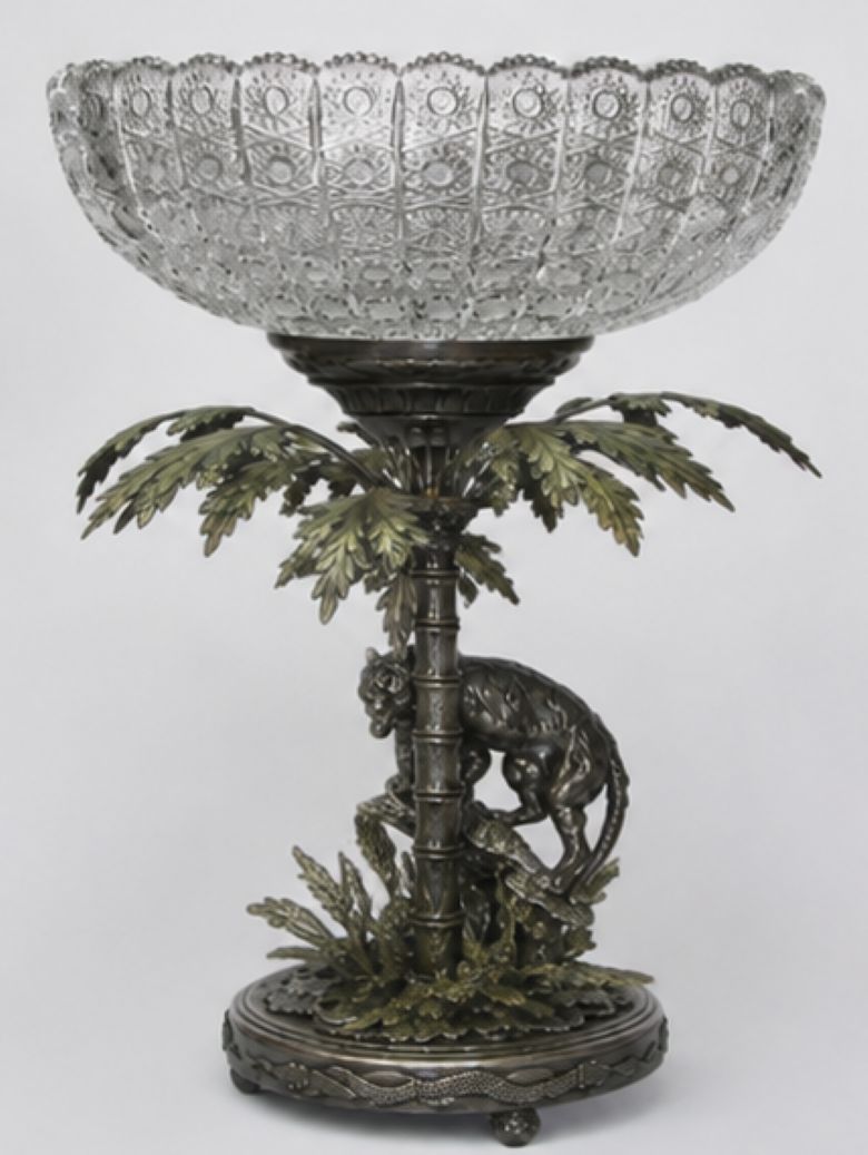 An ornate pedestal bowl featuring a clear cut-glass bowl mounted on a bronze and silver-toned metal base sculpted with a palm tree and a leopard, set against a plain light gray background.
