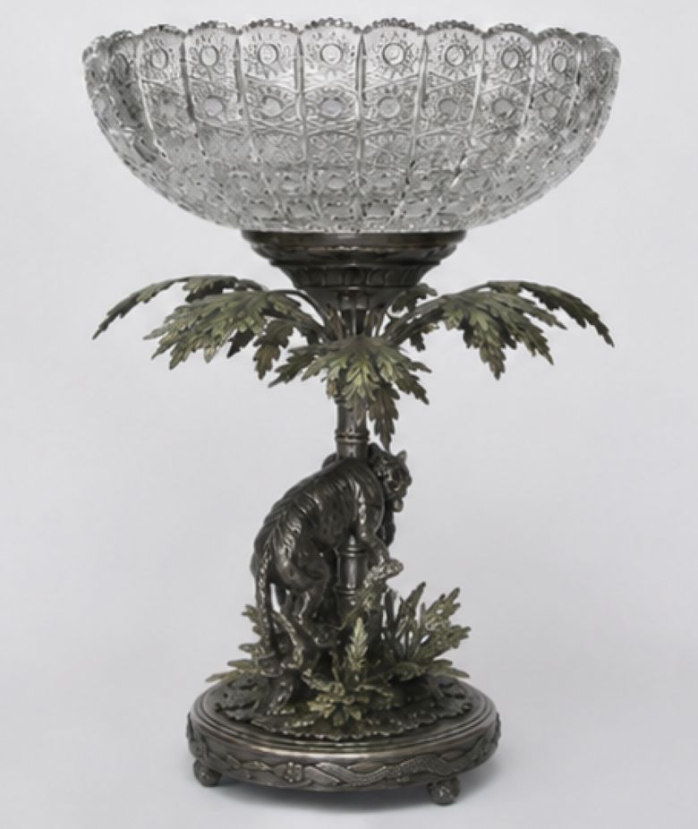 An ornate pedestal bowl featuring a clear cut-glass or crystal bowl mounted on a dark bronze or pewter base sculpted with a palm tree and a seated figure, photographed against a light gray studio background.