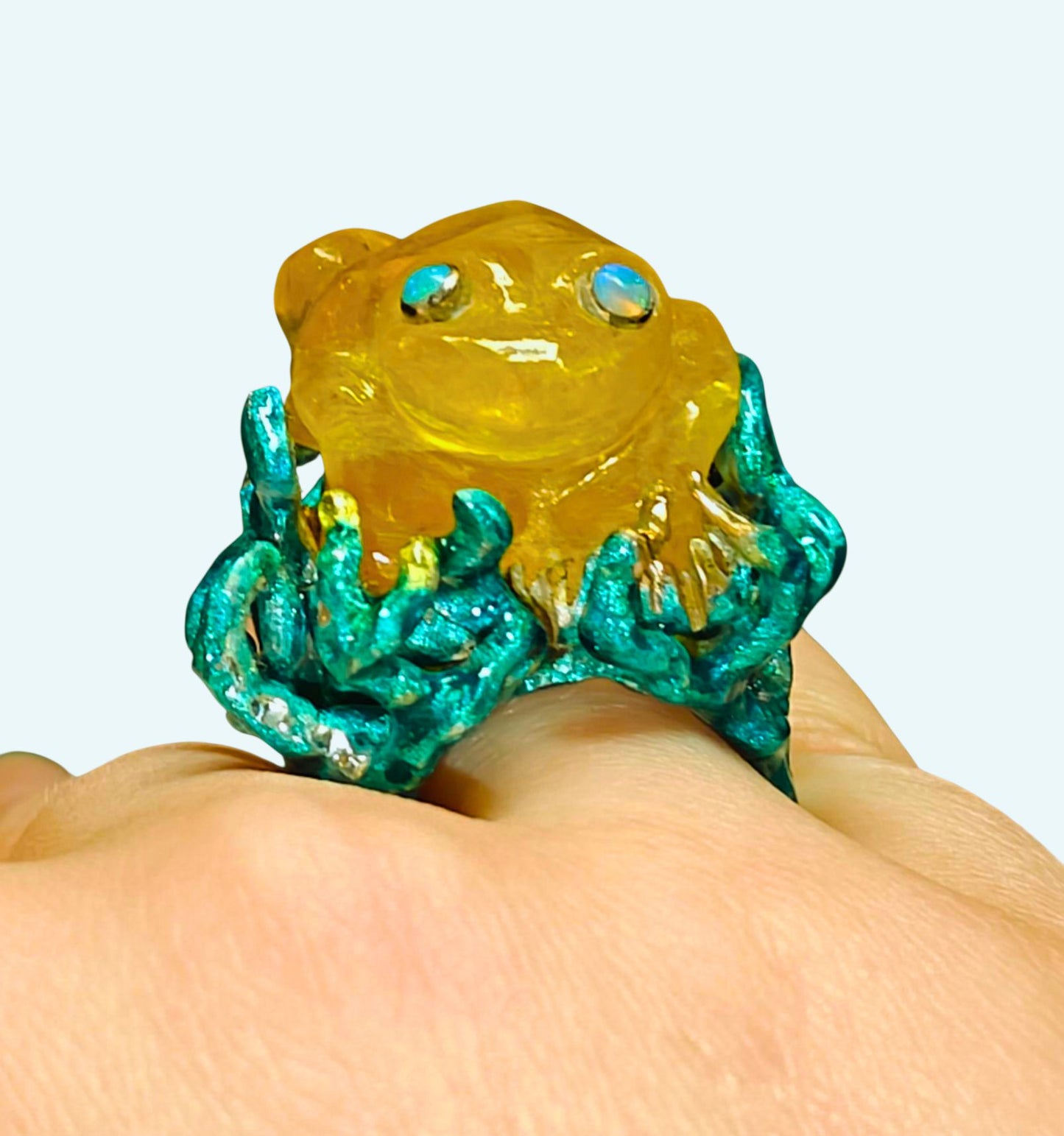 Kelp Dancer ring on hand, handcrafted in 960 silver with a 100 ct yellow lepidolite gemstone carved into a frog, detailed opal eyes, and vivid Limoges-style hand-painted enamel from Konforti Jewelry’s Stone Garden Collection.