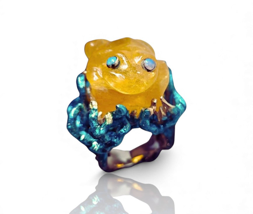 Angled image of the Kelp Dancer ring showcasing the carved 100ct lepidolite frog, shimmering opal eyes, and vivid layered enamel artwork on the 960 silver setting, evoking the movement of underwater kelp.