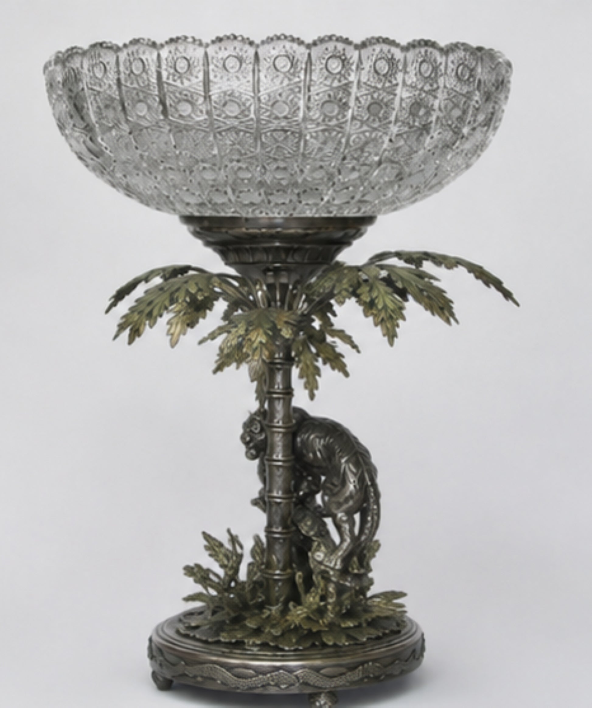 An ornate pedestal bowl featuring a clear cut-glass bowl mounted on a bronze and green metal base sculpted with a palm tree and a lion.