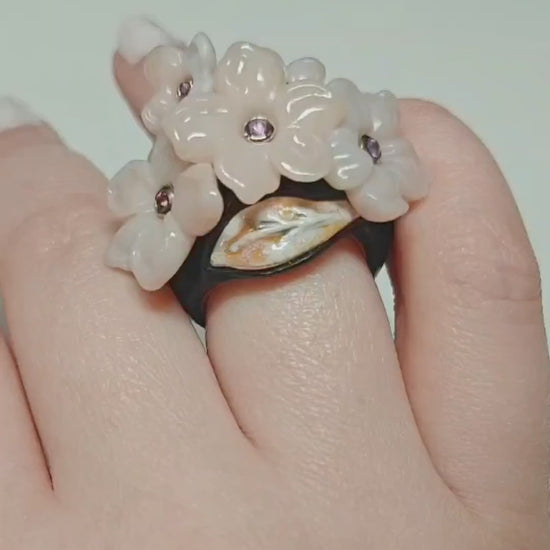 On-hand video of the Pink Sakura ring, highlighting the hand-carved pink opal flowers set with 0.2 ct pink sapphires, layered hot enamel over 18K gold, 14K pink gold inner band, and ebony wood outer base from Konforti Jewelry’s Stone Garden Collection.
