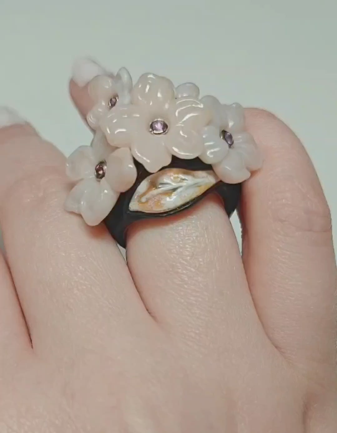 On-hand video of the Pink Sakura ring, highlighting the hand-carved pink opal flowers set with 0.2 ct pink sapphires, layered hot enamel over 18K gold, 14K pink gold inner band, and ebony wood outer base from Konforti Jewelry’s Stone Garden Collection.