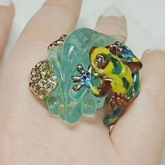 Video of the Summer Pond ring worn on the hand, featuring a hand-carved 64ct aquamarine gemstone in 18K yellow gold. The clip shows the gemstone’s depth, the vivid hot enamel painting inspired by Limoges techniques, and the 4.5ct multicolor sapphires that enhance the ring’s brilliance as it moves in the light.
