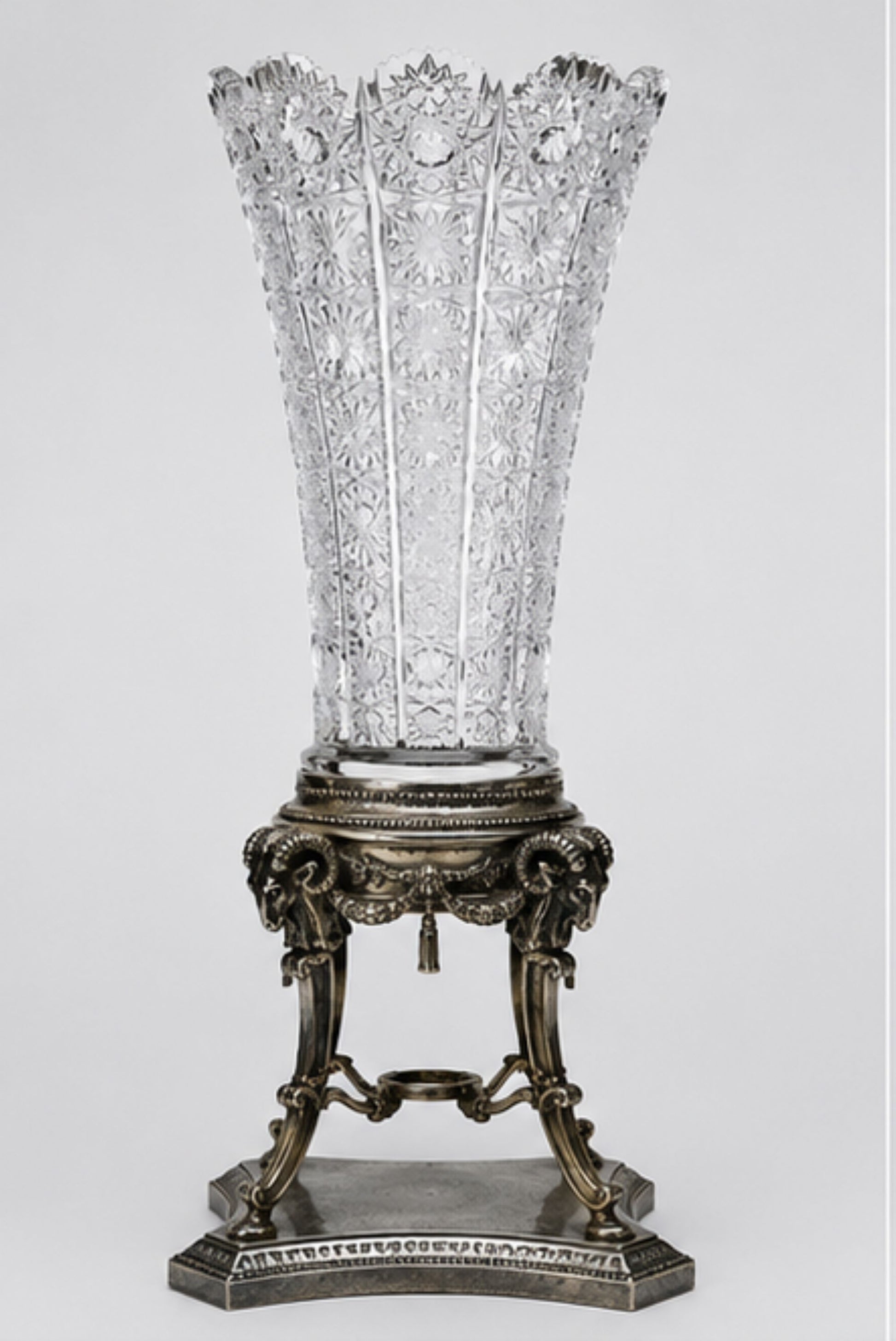 An ornate clear cut crystal vase with a flared, scalloped rim, displayed on an antique bronze-finished decorative pedestal stand against a light gray studio background.