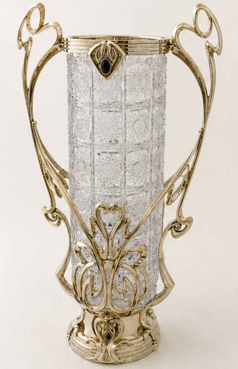 An ornate art nouveau style glass vase with a textured clear glass cylinder body and elaborate polished gold-toned metal handles and base, set against a plain light background.