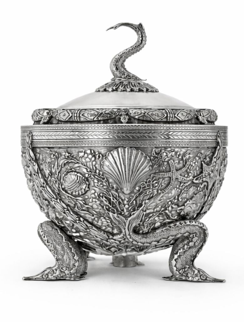 An ornate silver-toned lidded container or candelabra with intricate relief carvings of sea creatures, shells, and floral motifs, standing on three sculpted legs against a plain white background.