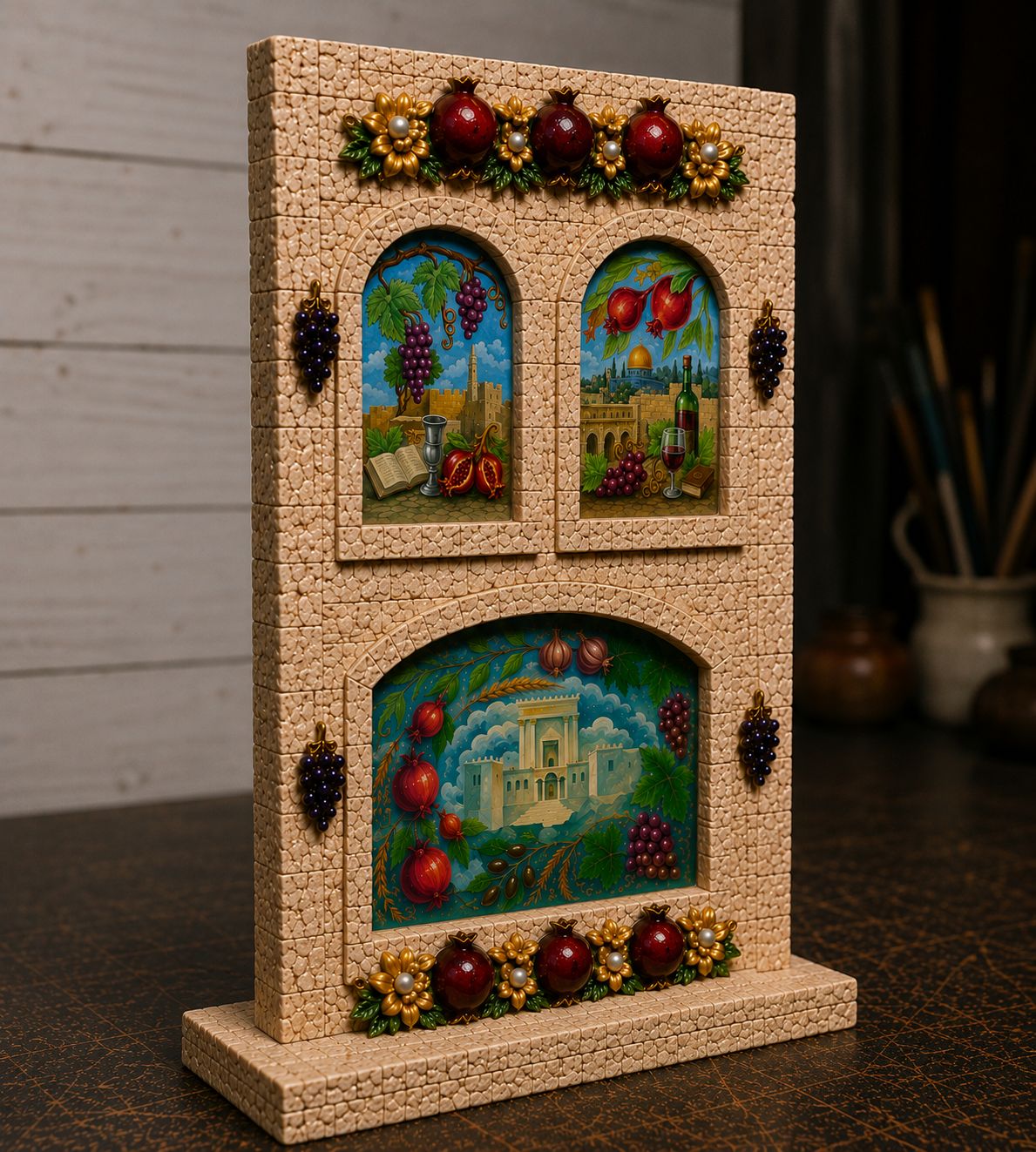 A decorative freestanding tabletop sculpture or plaque with a beige stone-textured frame, featuring three arched inset scenes with grapes, pomegranates, and architectural details, plus hanging dark berry motifs and floral accents, shown on a dark tabletop against a softly blurred indoor background.