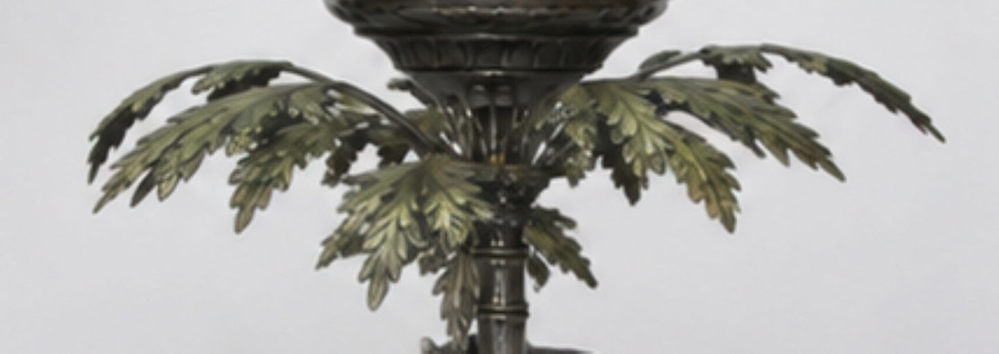 An ornate bronze metal candle holder or lamp base featuring a detailed leafy palm frond design around a central stem, set against a plain light gray background.