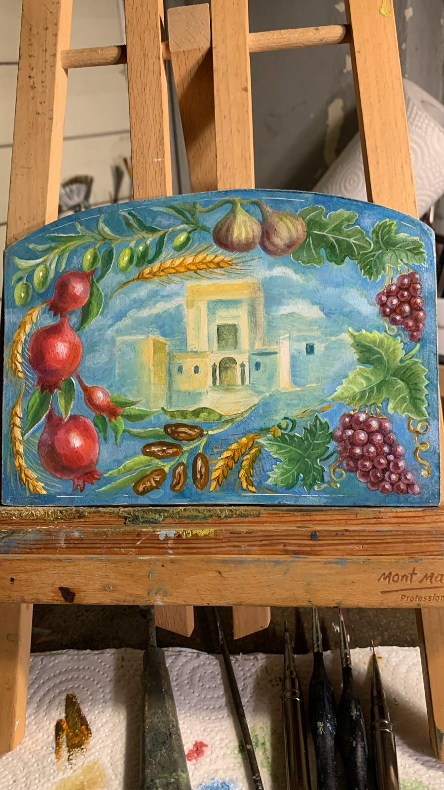 A hand-painted wooden panel depicting a Mediterranean village scene with pomegranates, grapes, wheat, and garlic, displayed on a wooden artist's easel.