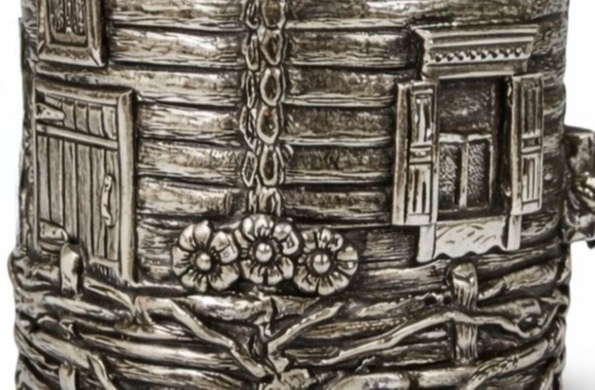 Close-up of sterling silver cup holder showing detailed cottage window, shutters, lantern, wood log textures, and handcrafted rural village scene